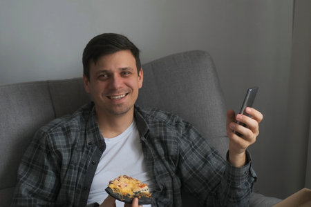 Portrait of young man at home with slice of pizza and smartphone in hands looking at camera and smiling sitting on sofa. He is happy, laughing. Wearing in shirt and t-shirt.の写真素材