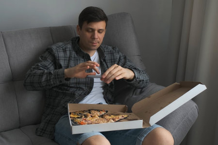 Young man is taking photos of pizza in box on his knees using smartphone. Unusual pizza with black dough. He is sitting on sofa at home, food blogger, social media life concept.の写真素材
