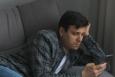 Man is browsing mobile phone typing message lying on couch at home. Young man is reading something in smartphone, working. Happy caucasian man chatting in social network, online web surfing internet.の写真素材