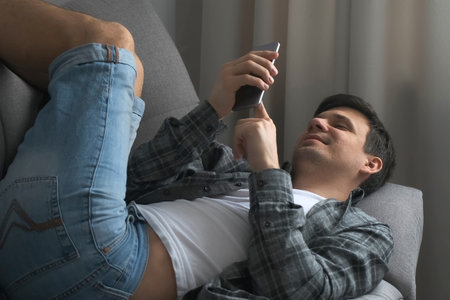 Young man is browsing smartphone lying on sofa finding a woman on dating site online. He is showing no and yes gestures and smiling. Virtual life of millennial man. Swipes pages on phone.の写真素材