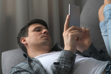 Young man is browsing smartphone lying on sofa finding a woman on dating site online. He is showing no and yes gestures and smiling. Virtual life of millennial man. Swipes pages on phone.の写真素材