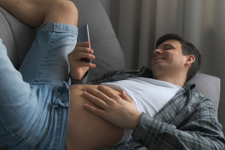 Man is browsing smartphone lying on sofa at home with exposed tummy. He is resting and scratching his belly. Looking on mobile phone screen in his free leisure time. Laughing and smiling.の写真素材