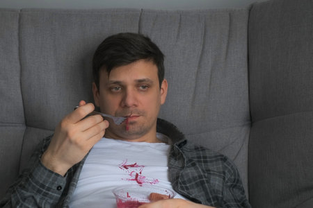 Man is eating beetroot salad from plastic container and falling it on white t-shirt sitting on the couch at home. He is in plaid shirt and white t-shirt.の写真素材