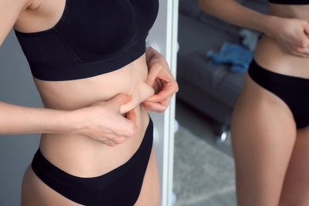 Woman in black underwear looking on her belly in mirror, close-up of her waist. Skin care and weight loss concept.の写真素材