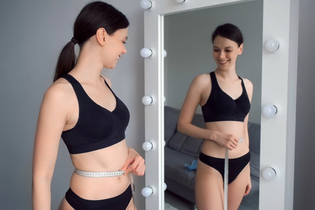 Young slim woman in black underwear measures her body with tape measure in front of the mirror. Concept of weight loss or obesity, healthy lifestyle. She is measuring her waist.の写真素材