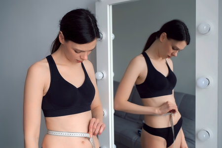 Young woman measures her waist looking at mirror. Concept of weight loss or obesity, healthy lifestyle. She is measuring her body in black underwear.の写真素材