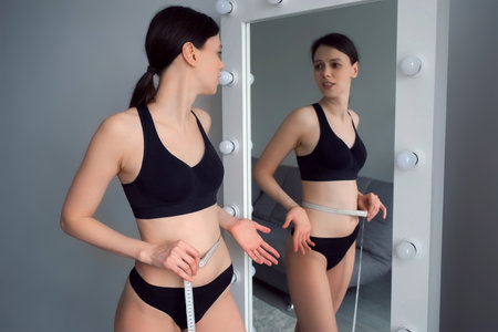 Brunette woman in black underwear measures her waist looking at mirror. Concept of weight loss or obesity, healthy lifestyle. She is measuring her body after losing weight.の写真素材