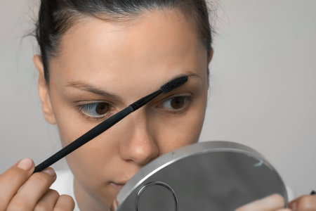 Woman is brushing her eyebrows before painting to give them the necessary shape looking at mirror. Preparing to eyebrow paint beauty procedure for herself at home. Female without makeup.の写真素材