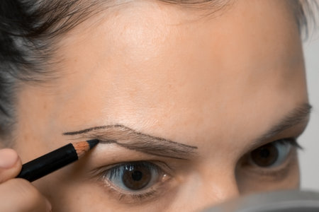 Young woman painting contour of eyebrows using pencil before painting them, closeup. She is looking at mirror. Preparing to eyebrow paint beauty procedure for herself at home. Female without makeup.の写真素材