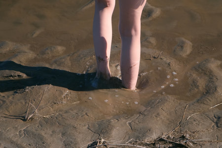 Boy is playing in wet mud sand on riverbank, barefooted feet closeup. He sinks deep into the sand with his feet and gets out of there. Wild swampy area near the river.の写真素材