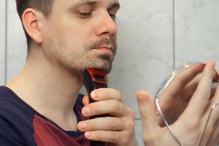 Portrait of young unshaven man shaving cheeks, beard, moustache with electric safety razor. He is looking at small mirror. takes care of hygiene. Shaves stubble with shaving machine in morning.の写真素材