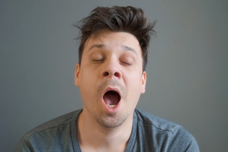 Portrait of young shaggy man with hangover is yawning looking at camera on grey background. Headache, human bad habits. Morning after a great party.の写真素材