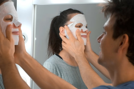 Man is applying a rejuvenating cosmetic textile mask on womans face from his face near the mirror at home. Concept of male and female beauty and skincare.の写真素材