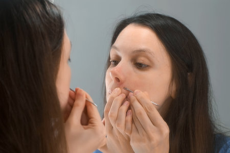 Young brunette woman squeezes out pimples on her face in front of the mirror at home. She is squeezing out acne using metal tool for it, needle with ring. She is cleaning her face.の写真素材