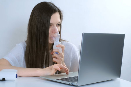 Sick young woman is using nebulizer and inhaler for treatment at home and working on laptop. She is breathing in mask. Fibrosis, covid-19, asthma, copd. Treatment, cure and procedure, therapy.の写真素材