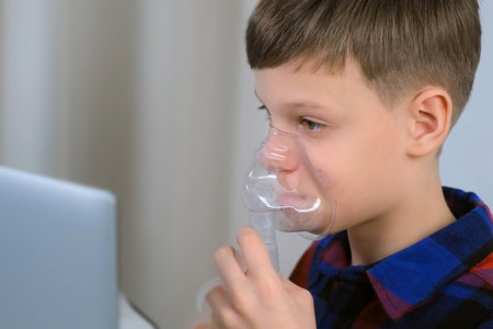 Sick tired boy is breathing in mask using nebulizer for inhalation at home and watching laptop, side view. Fibrosis, covid-19, asthma, copd. Treatment, cure and procedure. Using inhaler for therapy.の写真素材