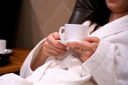 Cup of tea in womans hands drinking tea resting lying in armchair in spa sauna. female in white bathrobe enjoys weekend in spa salon. Rest and relax, healthy recreation in spa wellness concept.の写真素材
