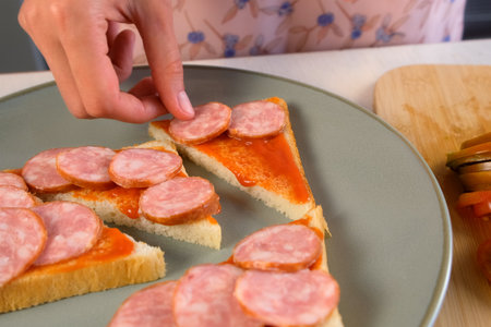 Woman putting smoked sausages slices on toasts bread with sauce cooking hot sandwiches on kitchen at home. Preparing snack fast food for herself. Tasty fresh meal for fast eating.の写真素材