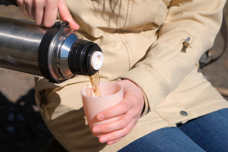 Traveler girl pouring tea from thermos to cup during hike sitting on bench in landscaped nature park or forest. Hands woman tourist pours hot drink in mug. Hiking camping tourism outdoors activity.の写真素材