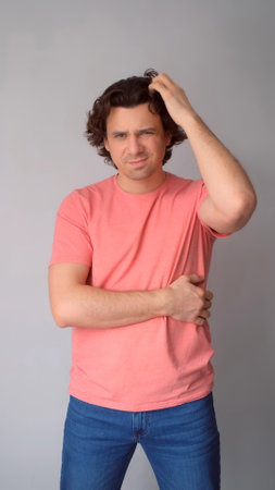 Confused man thinks scratching head by hand looking at camera on gray background. Curly middle aged guy feeling misunderstanding, trying to figure out situation. Pensive male wear jeans pink t-shirt.の写真素材
