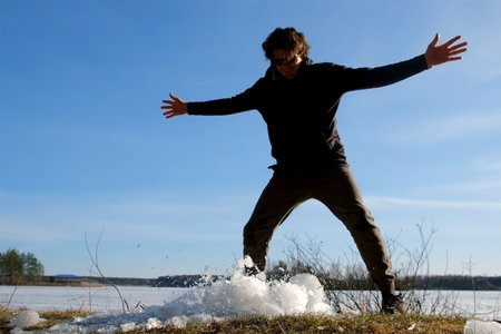 Playful man breaks a block of ice near frozen river on nature in sunny day. Guy throwing ice on the land to break it up. Large piece of ice breaks up into small fragments. Winter, early spring season.の写真素材