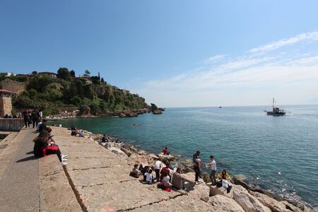 People sits along the coast and look at the Mediterranean sea. A tourist ship is sailing on the sea. Tourist port. Turkey, Antalya.のeditorial素材