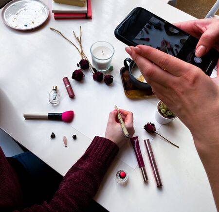 On the office desk is a sheet of white paper for inscription. Around laid out cosmetics, dry roses, jewelry, a cup of coffee and a cactus, books. Female hand holds a pen. Other hands take a photo picture on the phone. Flat lay. View from above.のeditorial素材