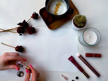 On the office desk is a sheet of white paper for inscription. Around laid out cosmetics, dried roses, jewelry, a cup of coffee and a cactus. Women's hands hold perfume. Flat lay. View from above.のeditorial素材