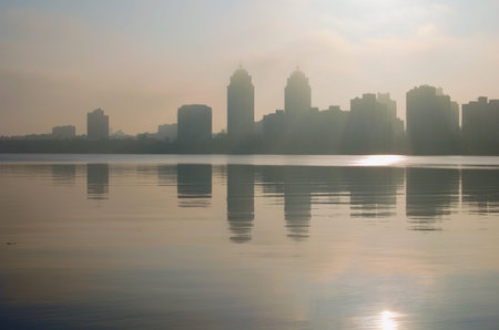 Abstract landscape view of black silhouettes of high-rise buildings in thick fog. Buildings reflected in tranquil water. Natural composition. Autumn in Kyiv, Ukraine.の写真素材