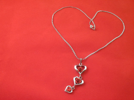 Silver chain and pendant with hearts, red roses and crystal. Lying in the form of heart on red backgroundの写真素材