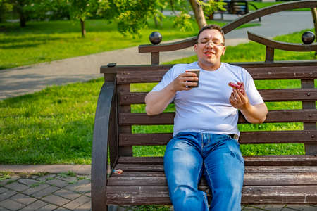 funny guy enjoys a donut with coffee sitting on a bench in the parkの写真素材