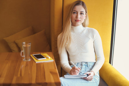 Portrait of a beautiful young blonde student girl with long hair sitting in cafe and studyingの写真素材