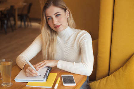 Portrait of a beautiful young blonde student girl with long hair sitting in cafe and studyingの写真素材
