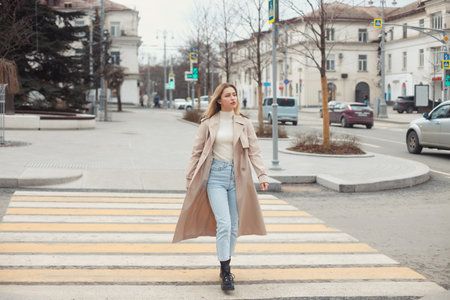 Young beautiful blonde hair woman in fashion clothes: beige trench coat, black boots and jeans crossing the road street.の写真素材