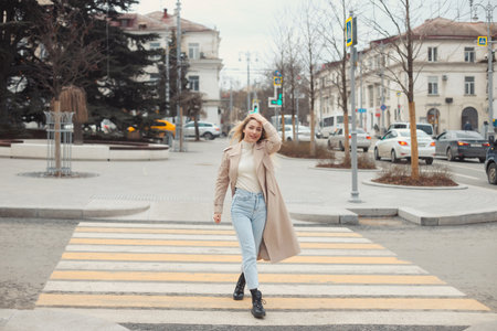 Young beautiful happy blonde hair woman in fashion clothes: beige trench coat, black boots and jeans crossing the road street.の写真素材