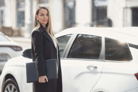 Street style portrait of serious business woman wearing white jacket and black trousers, walking along the city with laptop, gets into the carの写真素材