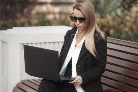 Street style portrait of beautiful business woman in sunglasses sitting on a bench with laptop