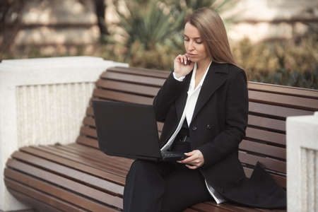 Street style portrait of beautiful business woman in black coat sitting on a bench with laptop and talking phoneの写真素材