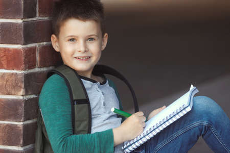 School boy sitting in the schoolyard against a brick wall doing homework and waits for parentsの写真素材