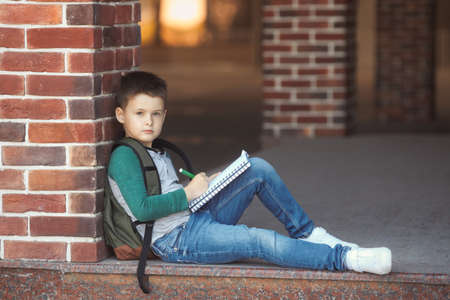 School boy sitting in the schoolyard against a brick wall doing homework and waits for parentsの写真素材