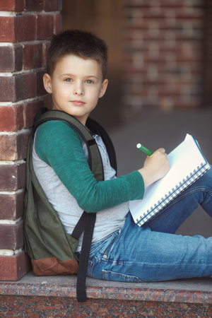 School boy sitting in the schoolyard against a brick wall doing homework and waits for parentsの写真素材