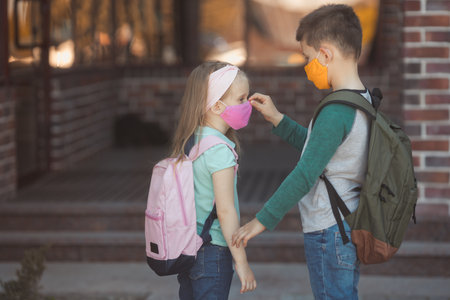 Brother takes care of little sister and correct the mask on her face at school. Cute kids wearing casual clothes, backpack and protective cotton mask, ready to study with a new normal covid-19.の写真素材