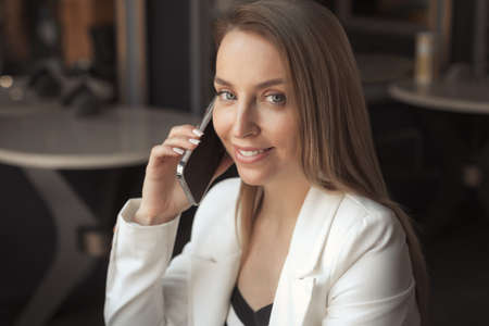 Portrait of smiling business woman with long blonde hair, talking on phone in cafe with dark black interiorの写真素材