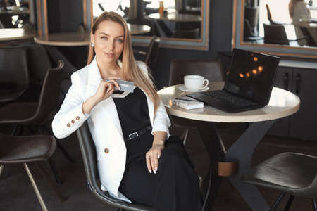 Business woman with long blonde hair drink coffee in a white blazer, holding credit card, working with laptop in cafe or doing online shoppingの写真素材