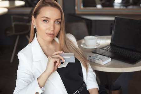 Business woman with long blonde hair drink coffee in a white blazer, holding credit card, working with laptop in cafe or doing online shoppingの写真素材