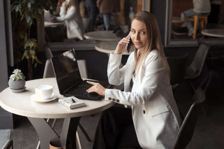 Business woman with long blonde hair drink coffee in a white blazer and working with laptop in cafe with dark black interiorの写真素材