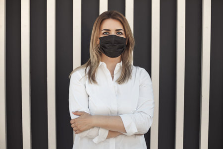 Closeup portrait of tired caucasian woman in black protective face mask during a coronavirus epidemic, standing near wall in white shirtの写真素材