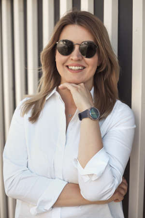 Lifestyle portrait of a smiling young confident woman 30-40 years old in a white shirt, wears sunglasses and a wrist watchの写真素材