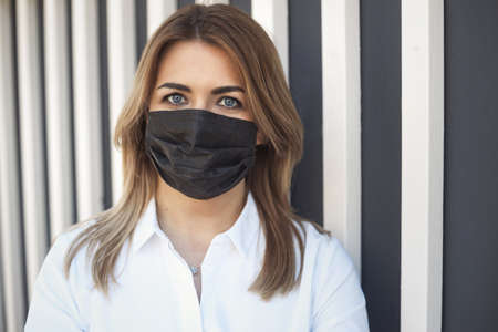 Closeup portrait of tired caucasian woman in black protective face mask during a coronavirus epidemic, standing near wall in white shirtの写真素材
