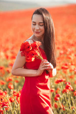 Young brunette woman 25-30 years old with long groomed hair in red dress enjoy a poppy flowers in fieldの写真素材